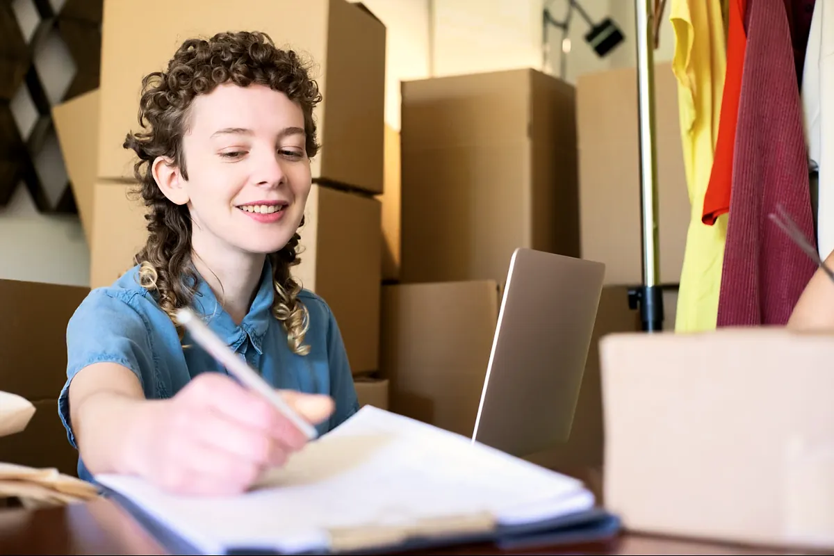 Shop owner smiling as she writes in a notebook beside stacked boxes of stock